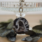 Achat Geode Schlüsselanhänger präsentiert vor Glasständer mit dunklen Blättern