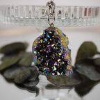 Aqua Aura Druse Schlüsselanhänger mit regenbogenfarbener Kristallfläche an Glasständer mit Blättern im Hintergrund