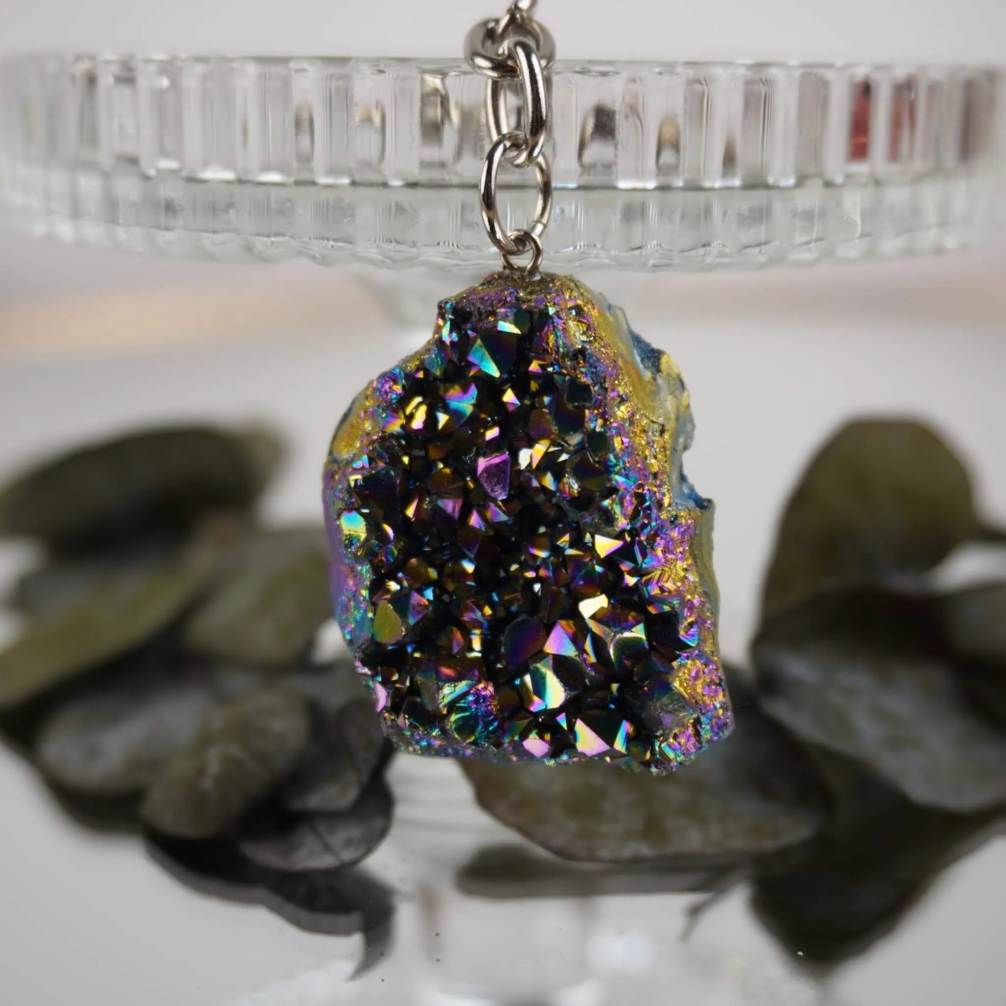 Aqua Aura Druse Schlüsselanhänger mit regenbogenfarbener Kristallfläche an Glasständer mit Blättern im Hintergrund