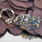 Regenbogenfarbener Aqua Aura Druse Schlüsselanhänger mit Spiegelung auf dunklen Blättern