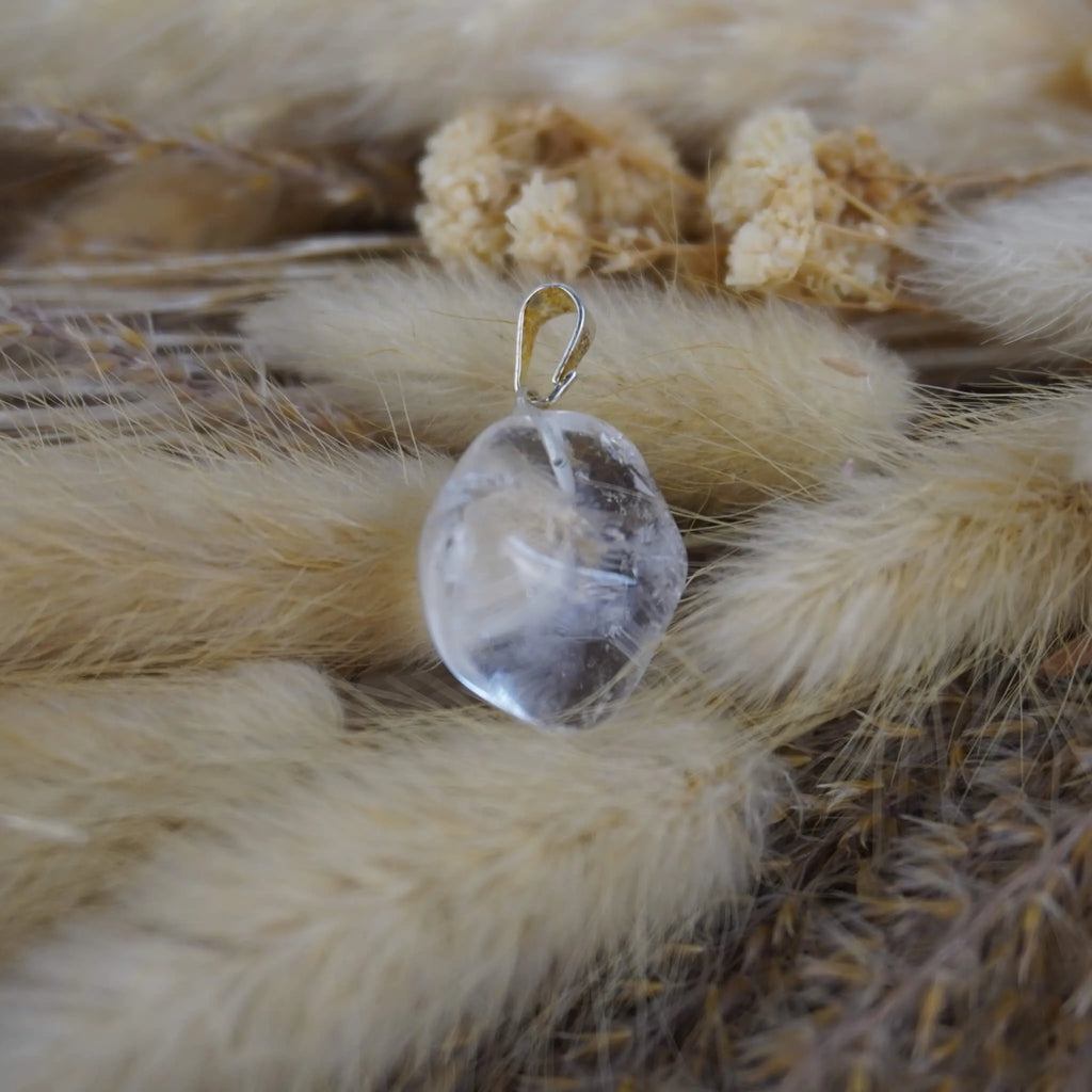 Bergkristall Trommelstein Kettenanhänger mit silberfarbener Öse dekoriert auf beigen Trockenpflanzen