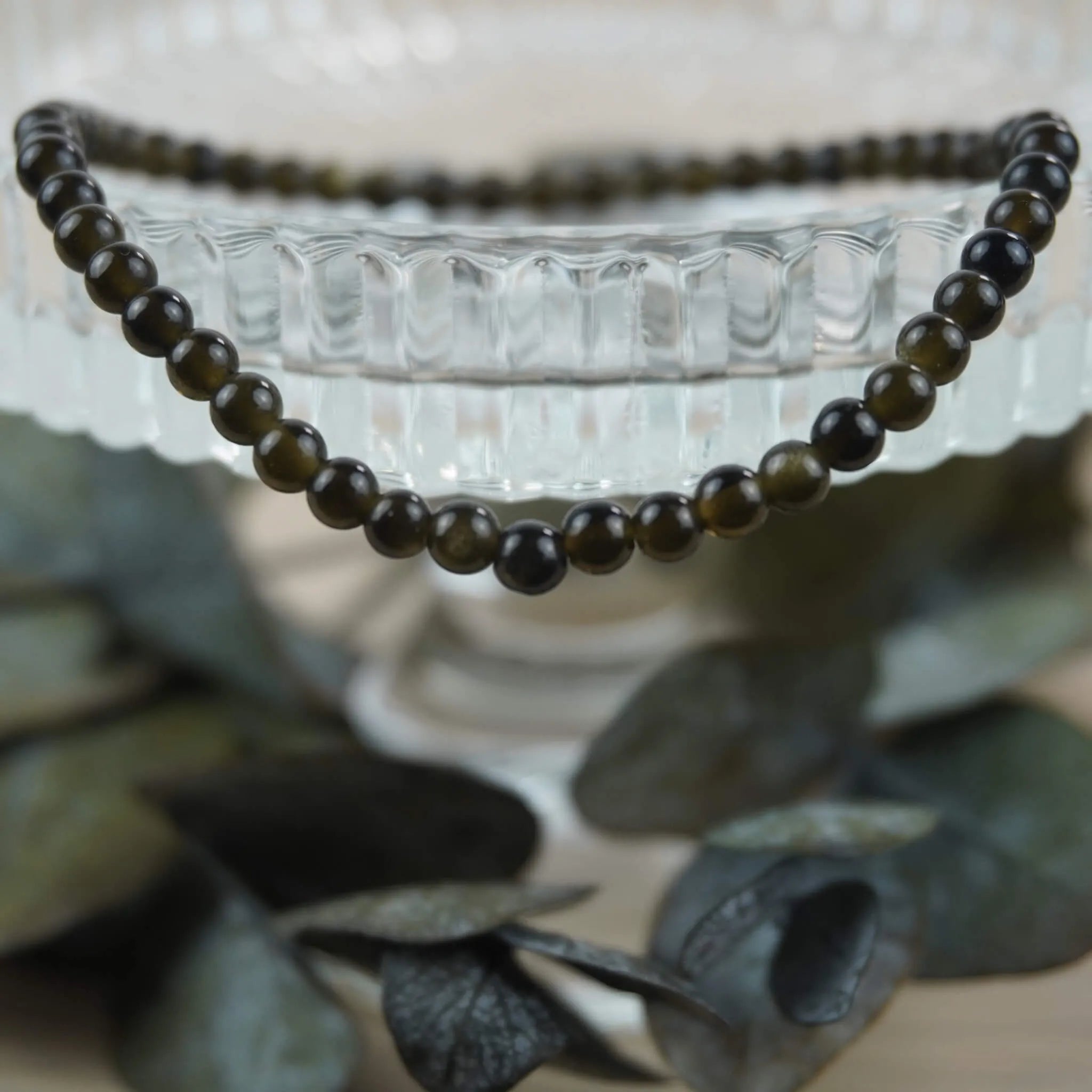 Goldenes Obsidian Kugelarmband auf Glas-Schale mit Eukalyptus stilvoll dekoriert