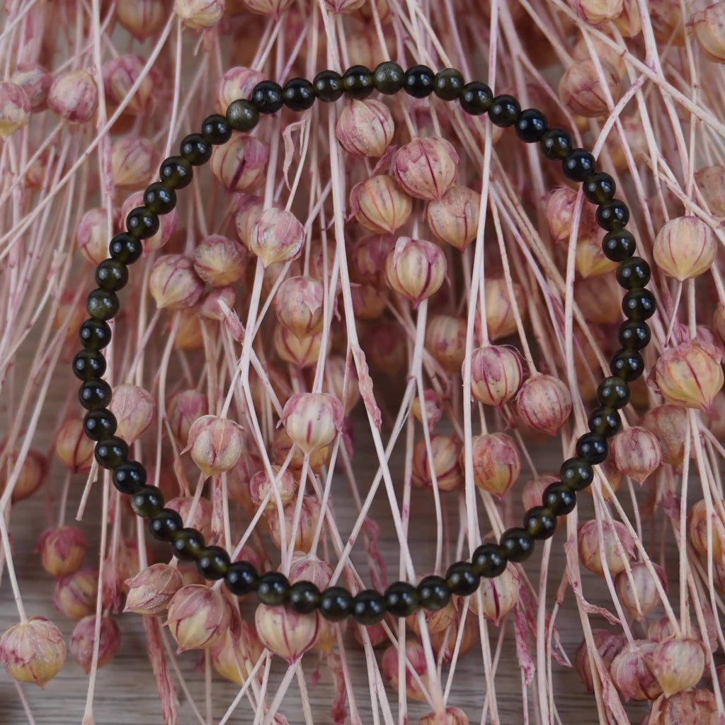 Goldenes Obsidian Kugelarmband vor rosafarbenen Trockenblumen mit warmem Kontrast