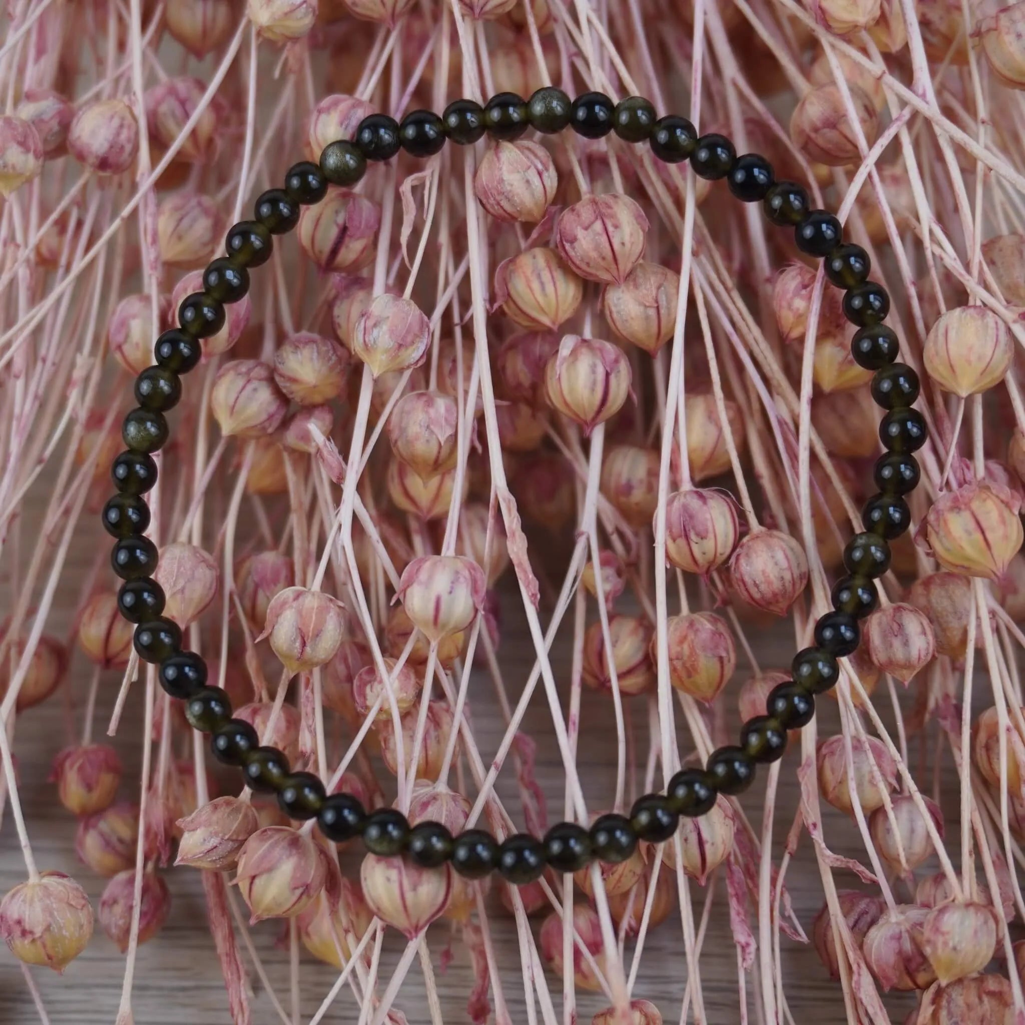Goldenes Obsidian Kugelarmband vor rosafarbenen Trockenblumen mit warmem Kontrast