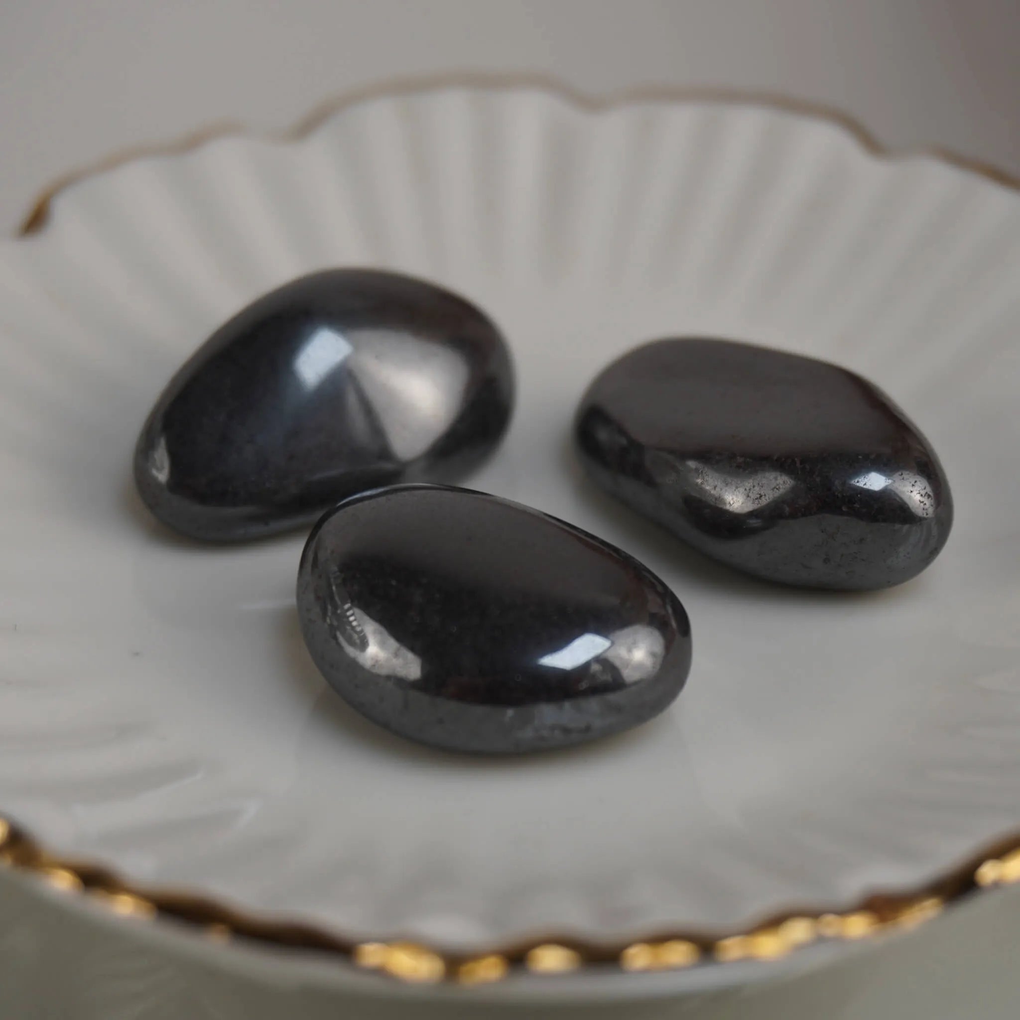 Drei polierte Hämatit Trommelsteine auf weißer Schale mit Goldrand