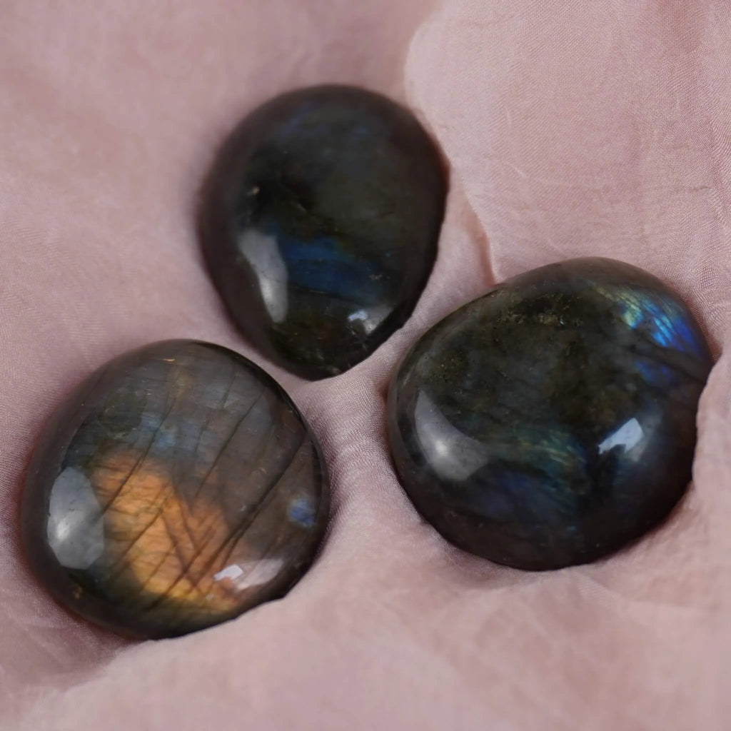 Drei Labradorit Trommelsteine mit blauem und goldenem Schimmer auf rosafarbenem Stoff