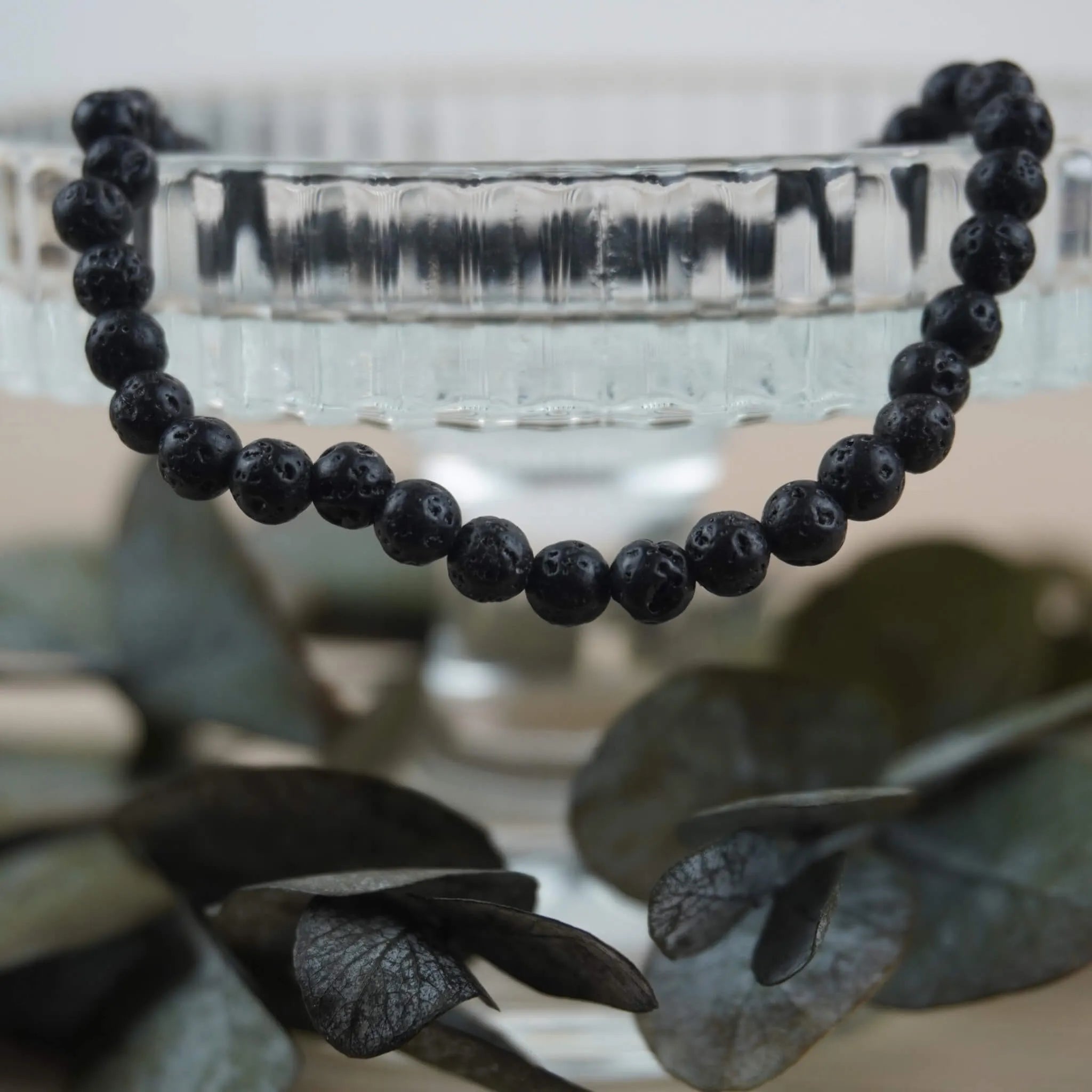Lavastein Kugelarmband an Glas-Schale mit dunklen Eukalyptusblättern arrangiert