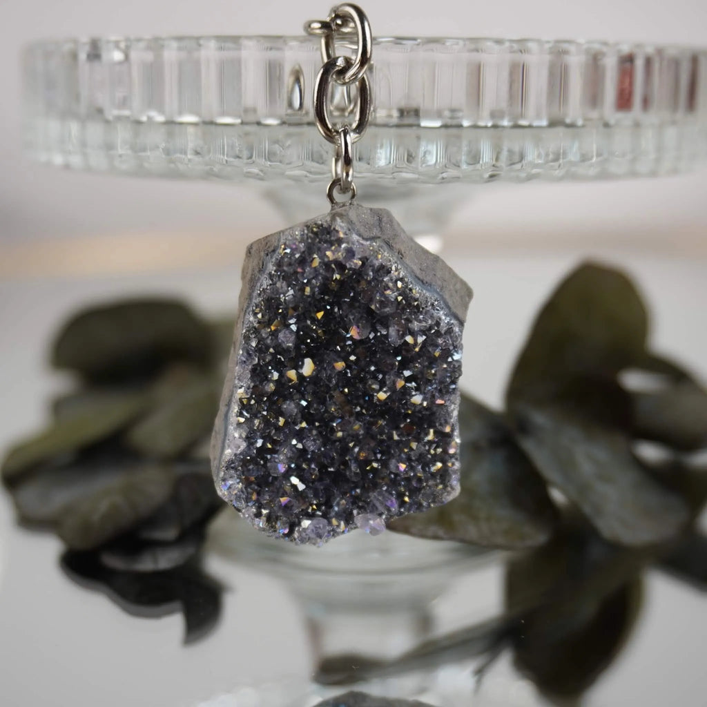 Lila Aqua Aura Druse Schlüsselanhänger arrangiert vor gläsernem Ständer und dunklen Blättern