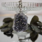 Lila Aqua Aura Druse Schlüsselanhänger arrangiert vor gläsernem Ständer und dunklen Blättern