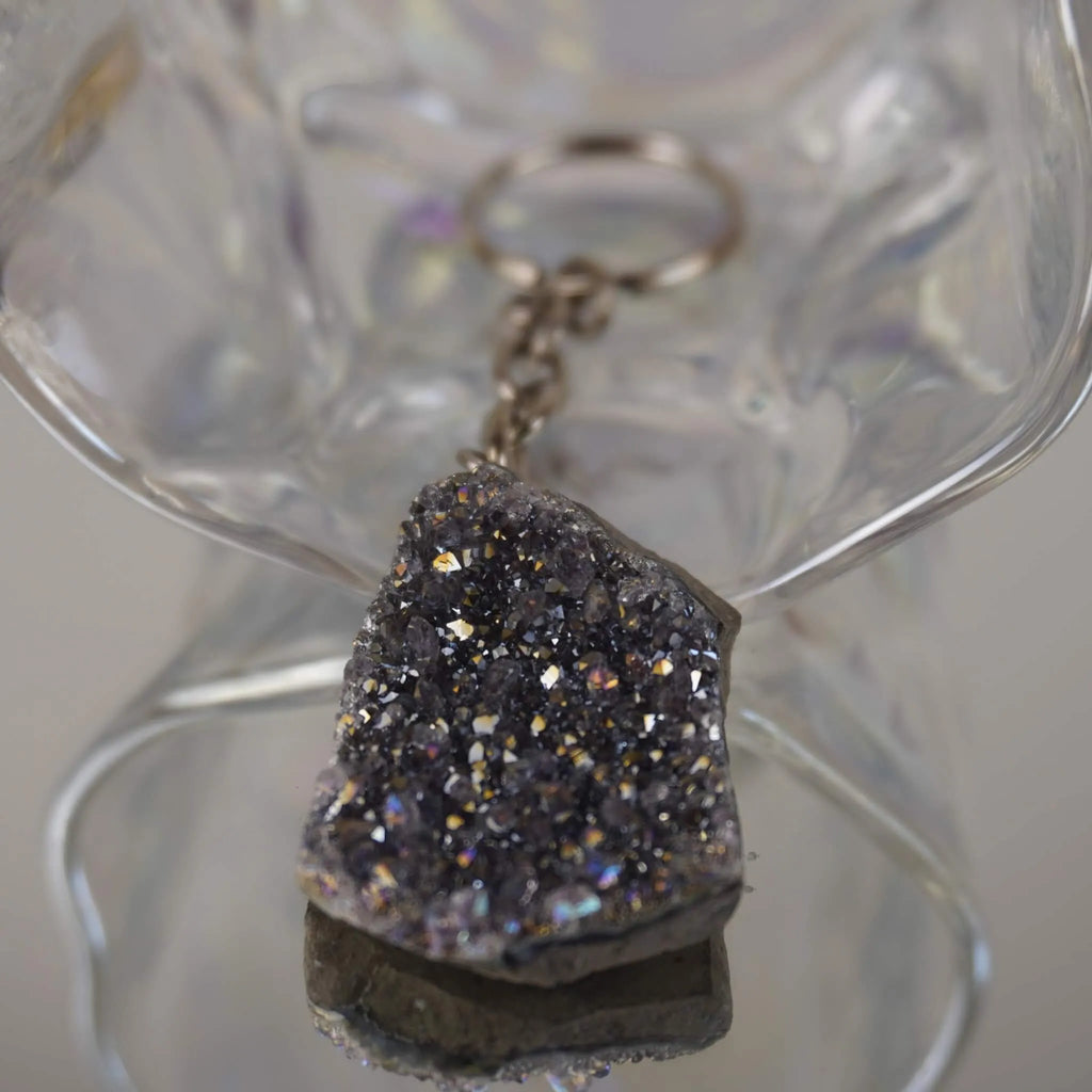 Lila Aqua Aura Druse Schlüsselanhänger mit funkelnder Kristallfläche vor transparenter Glasdeko