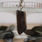 Rauchquarz Spitze Schlüsselanhänger vor Glasständer mit dunklen Blättern