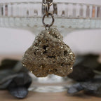 Roher Pyrit Schlüsselanhänger vor Glasständer mit dunklen Blättern