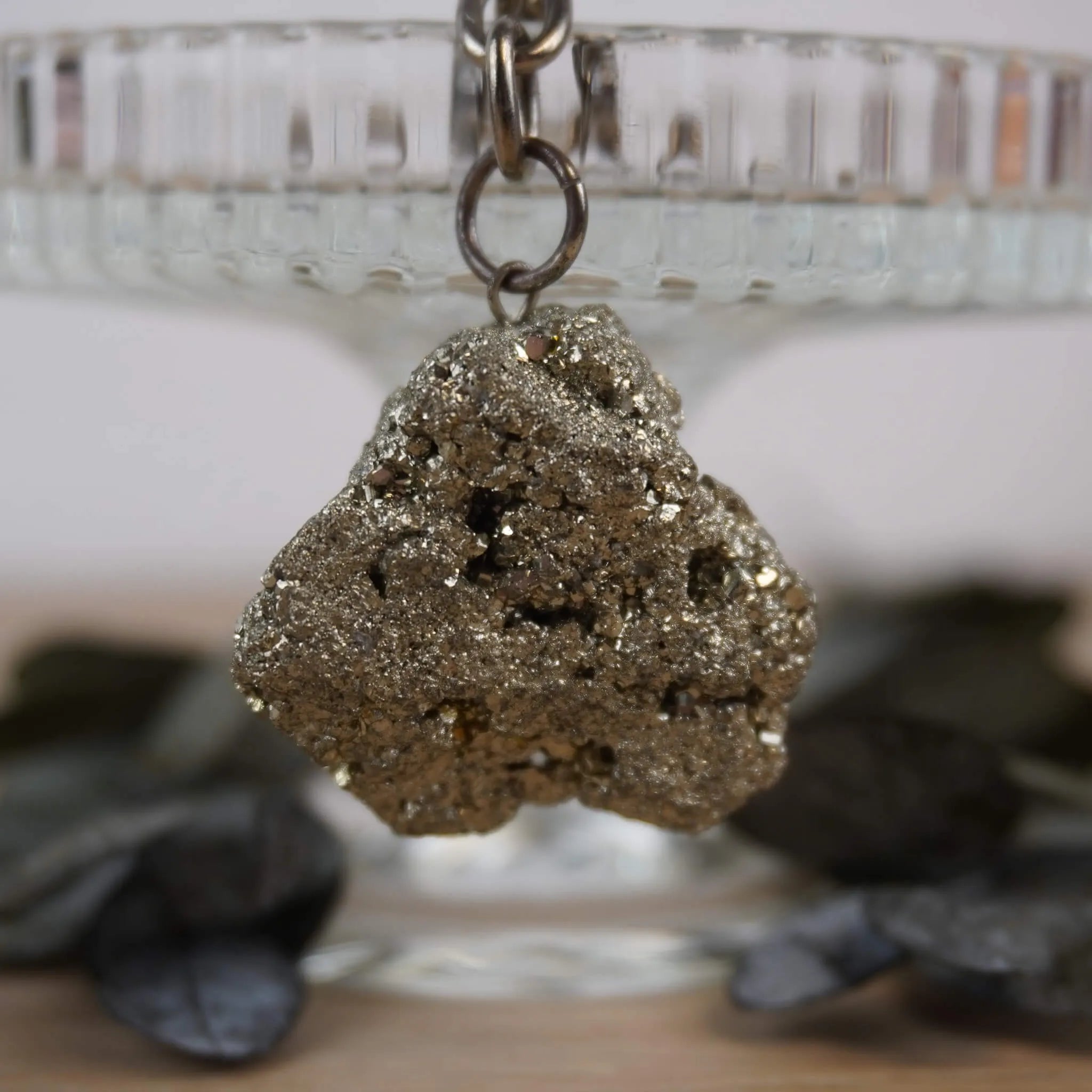 Roher Pyrit Schlüsselanhänger vor Glasständer mit dunklen Blättern