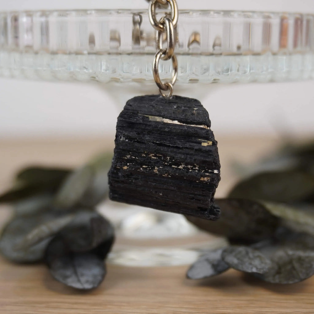 Roher schwarzer Turmalin Schlüsselanhänger vor Glasständer mit dunklen Blättern arrangiert