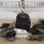 Roher schwarzer Turmalin Schlüsselanhänger vor Glasständer mit dunklen Blättern arrangiert