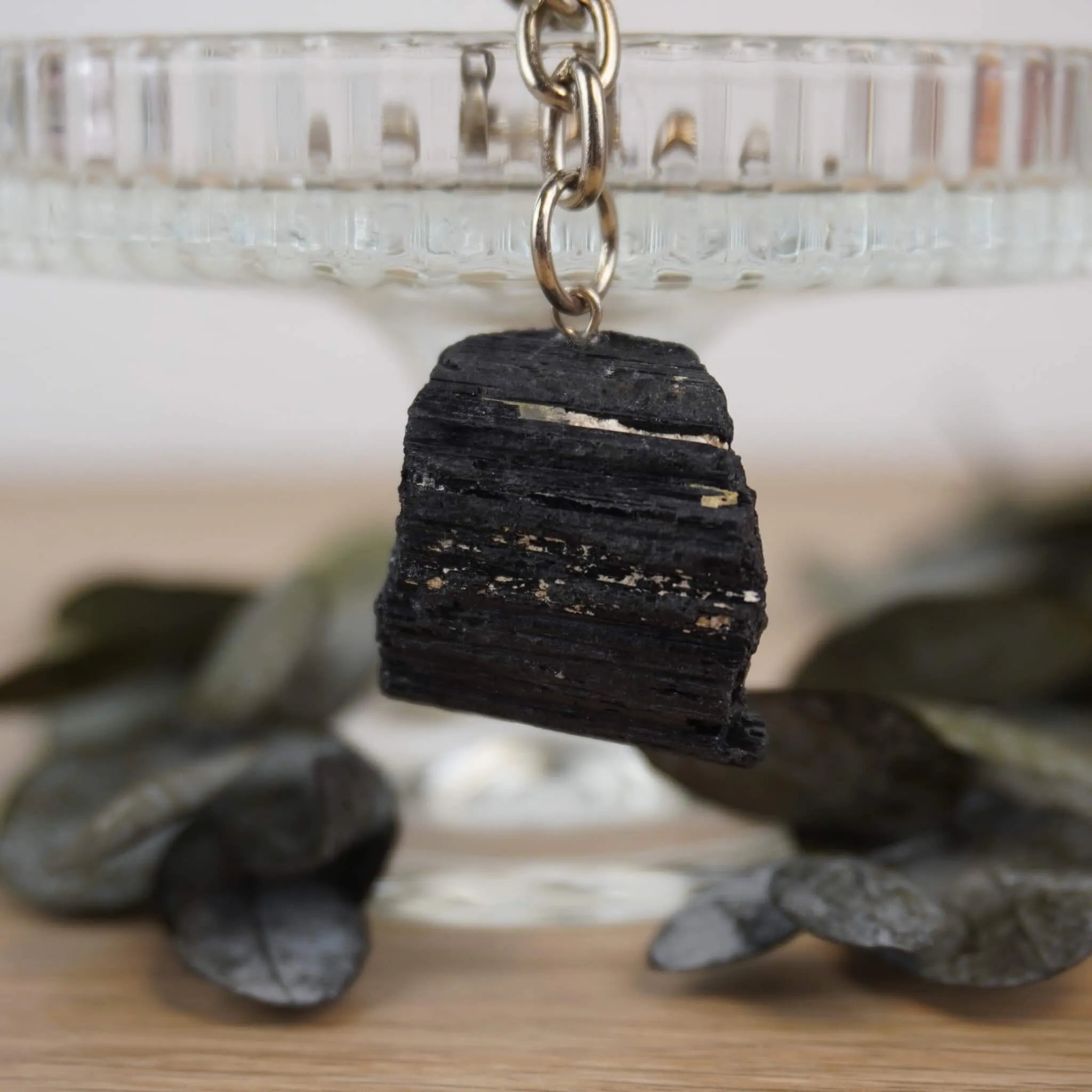 Roher schwarzer Turmalin Schlüsselanhänger vor Glasständer mit dunklen Blättern arrangiert