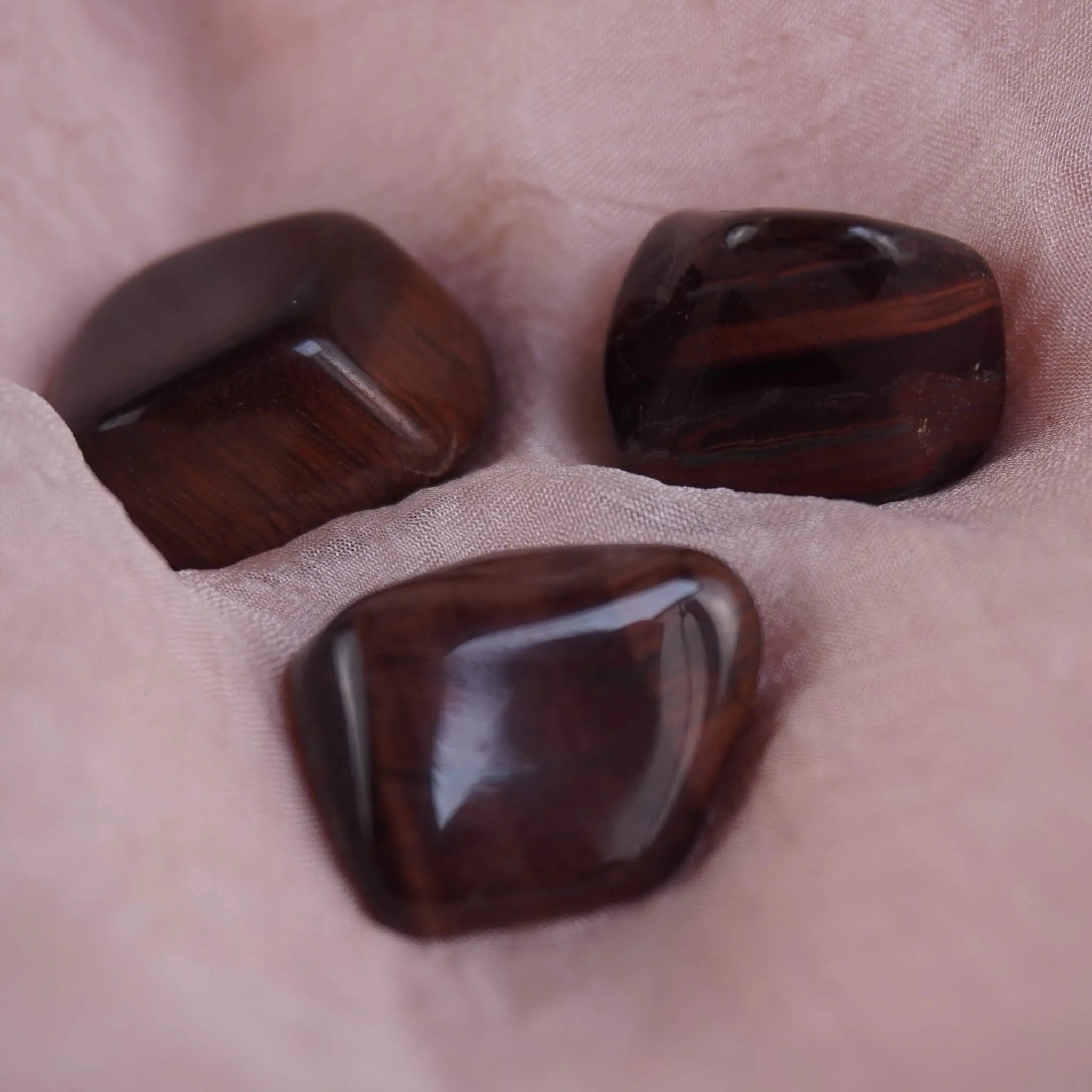 Drei dunkle rote Tigerauge Trommelsteine mit Glanz auf rosafarbenem Stoff