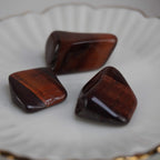 Drei polierte rote Tigerauge Trommelsteine auf weißer Schale mit Goldrand