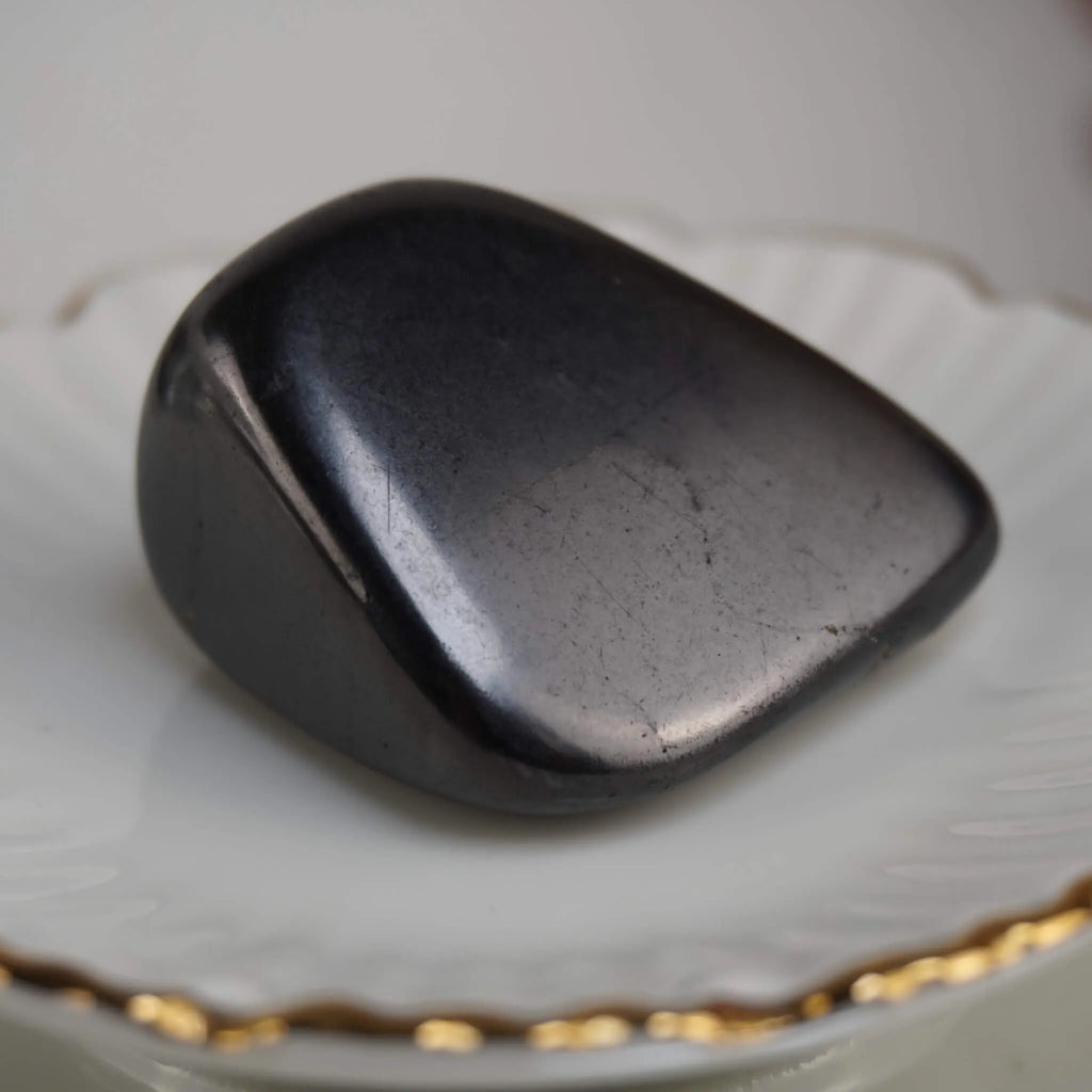 Polierter schwarzer Schungit Trommelstein auf weißer Schale mit Goldrand