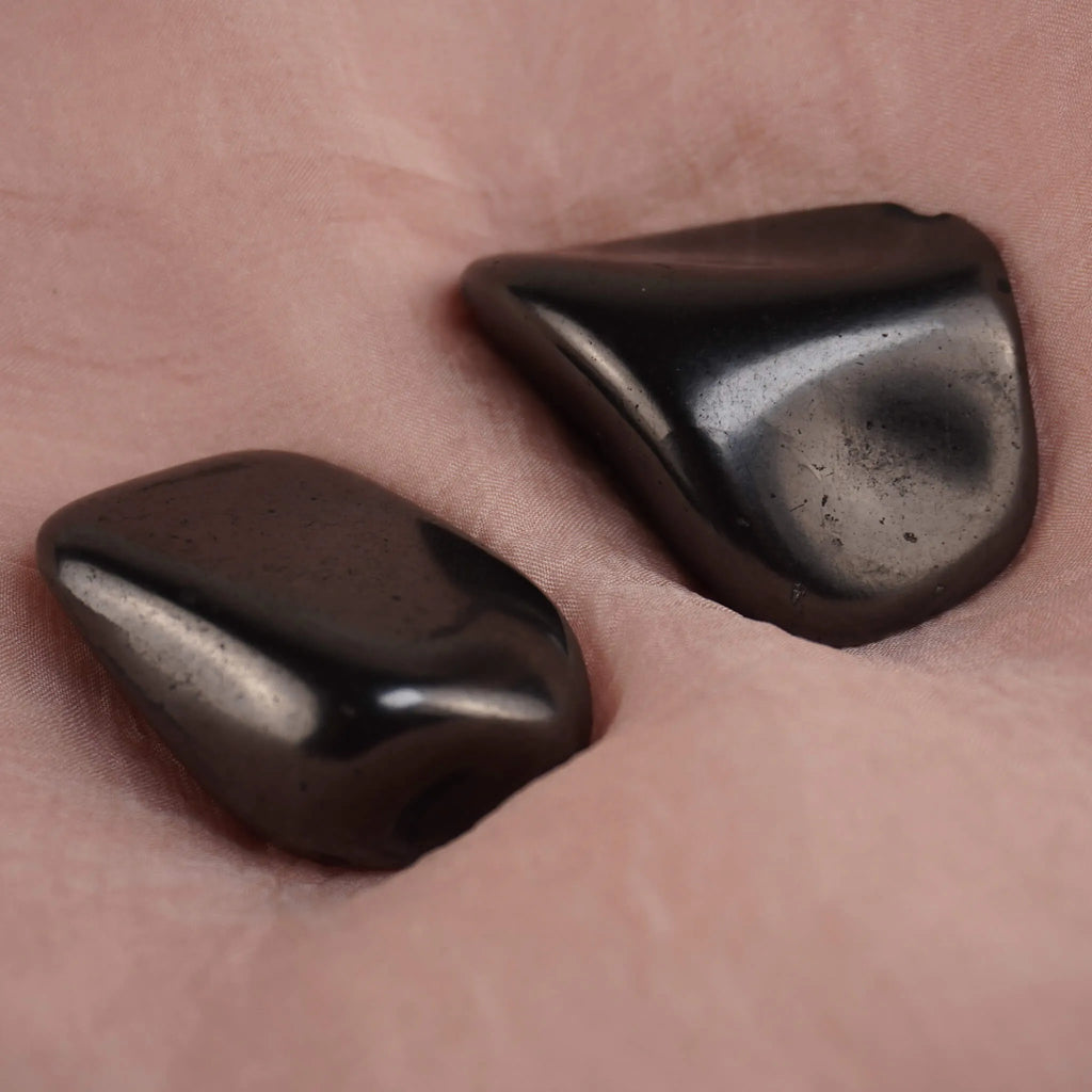 Zwei Schungit Trommelsteine präsentiert auf zart rosafarbenem Stoff