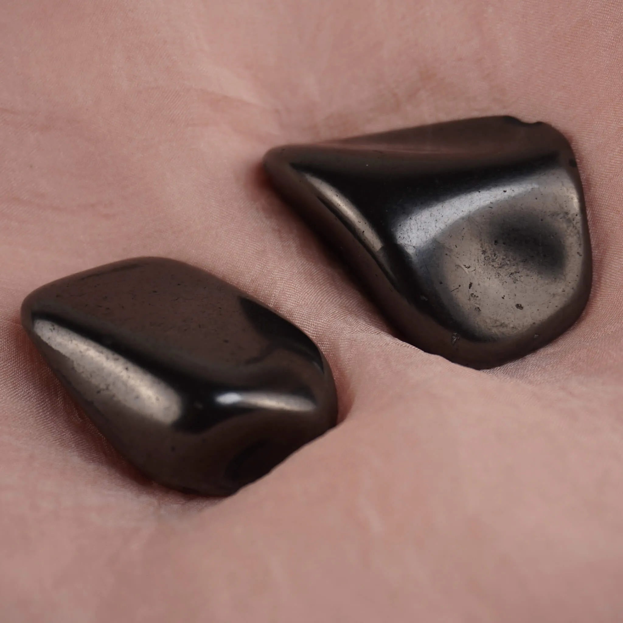 Zwei Schungit Trommelsteine präsentiert auf zart rosafarbenem Stoff