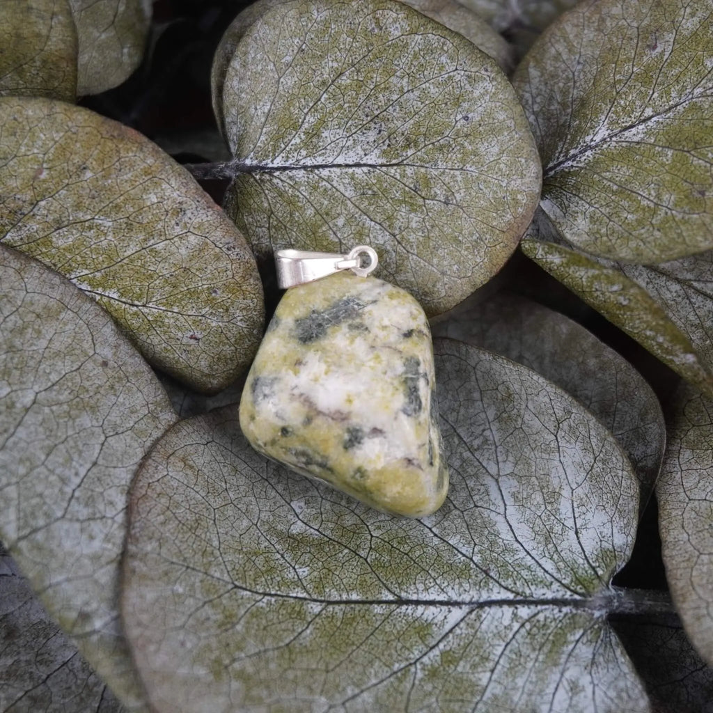 Serpentin Trommelstein Kettenanhänger mit silberfarbener Öse dekoriert auf grünen Eukalyptusblättern