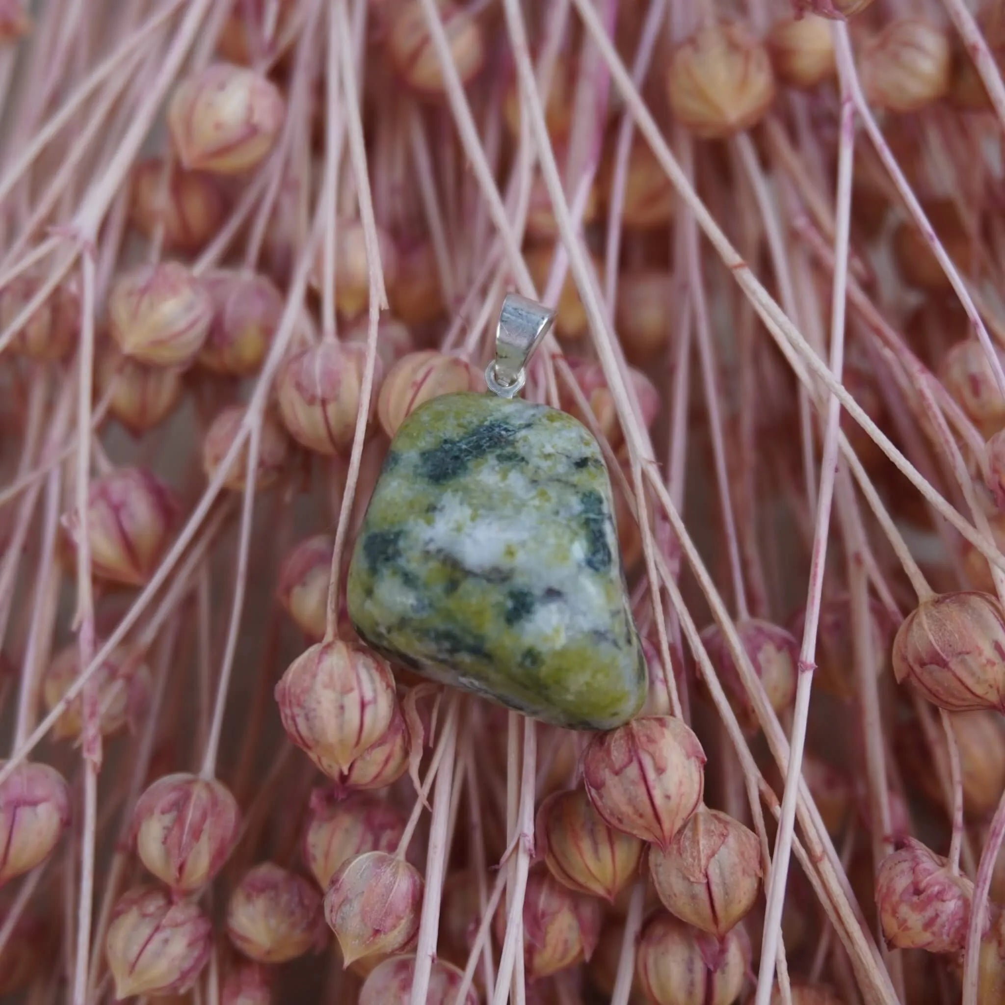 Serpentin Trommelstein Kettenanhänger mit silberfarbener Öse vor rosa Trockenblumen arrangiert