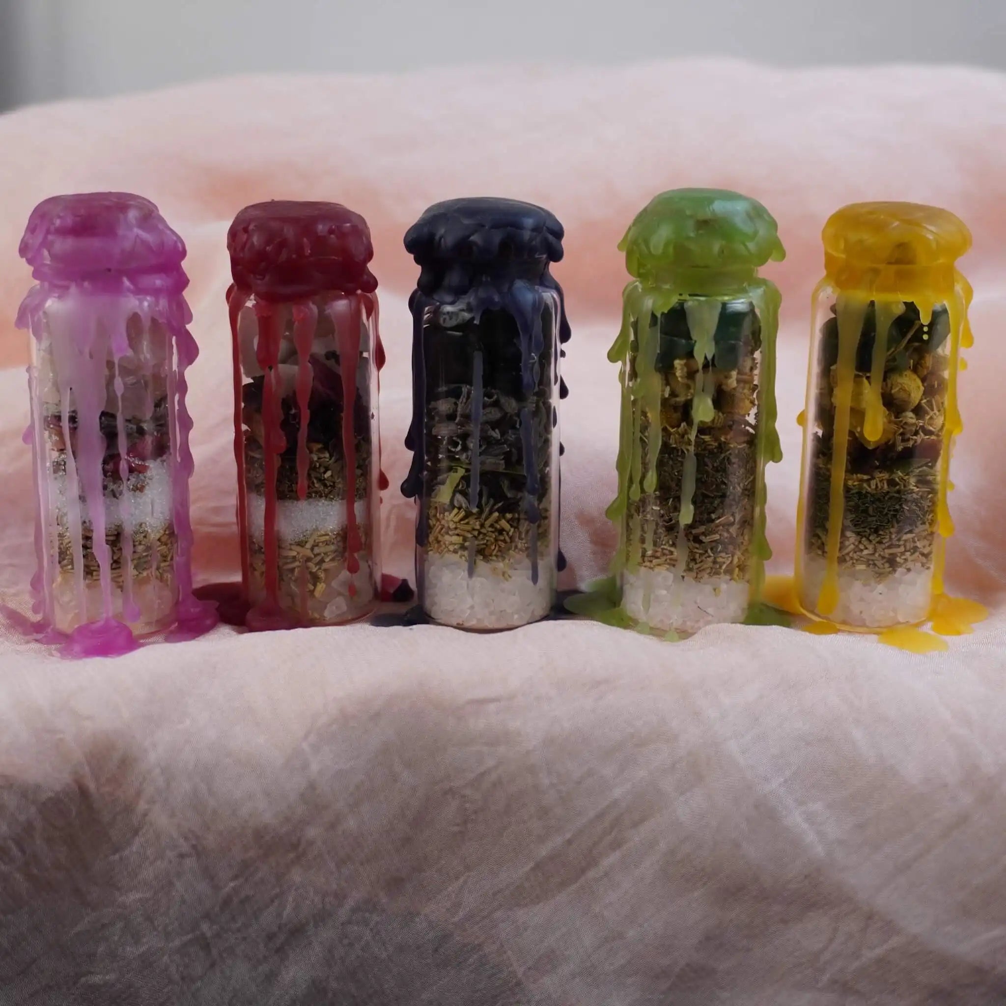 Handgemachtes Spell Jar Set mit fünf Gläsern, überzogen mit lila, rotem, blauem, grünem und gelbem Wachs auf rosa Stoff platziert